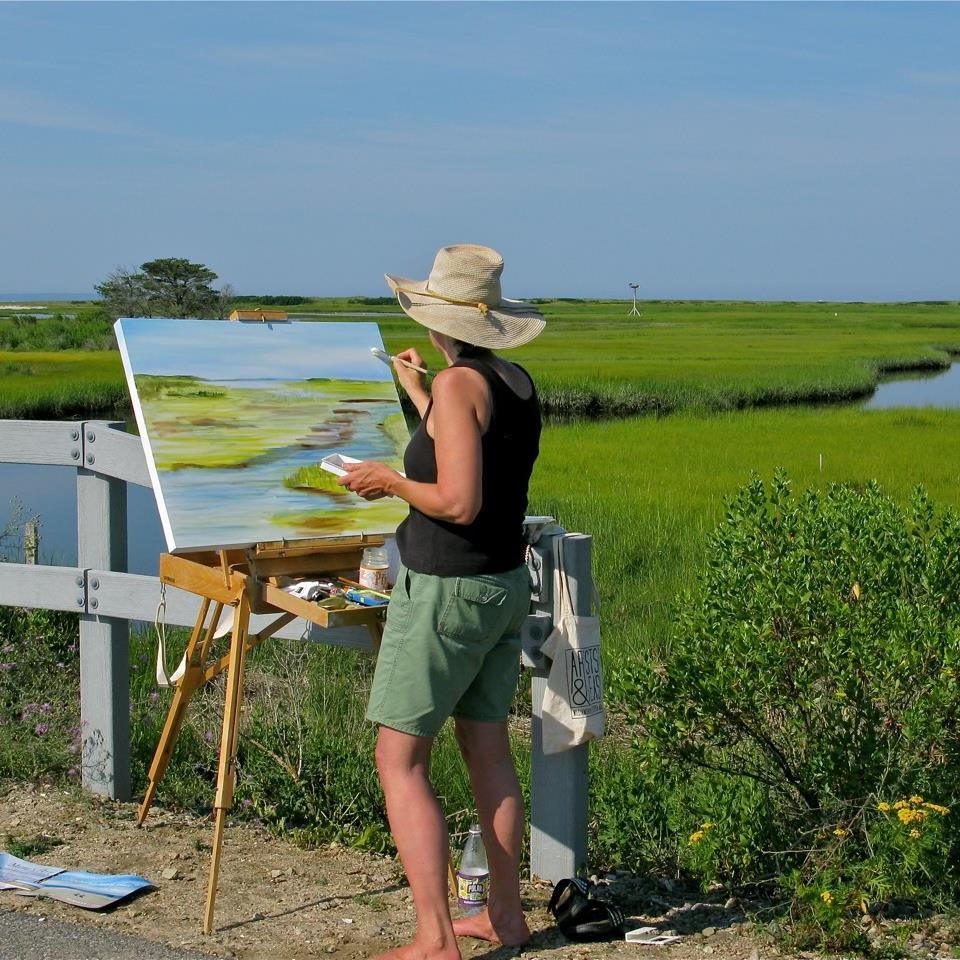 photo of artist Mimi Schlichter painting at an easel overlooking the beach