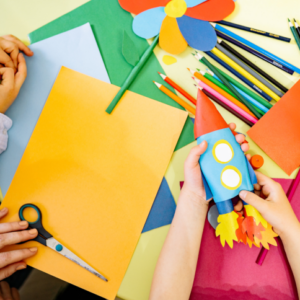 photo of three pale hands holding craft supplies and a handmade paper rocket