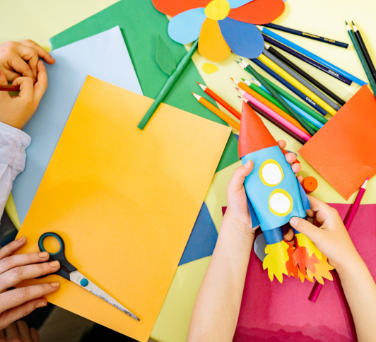 photo of three pale hands holding craft supplies and a handmade paper rocket